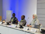 Three people sit in front of microphones at a board meeting.