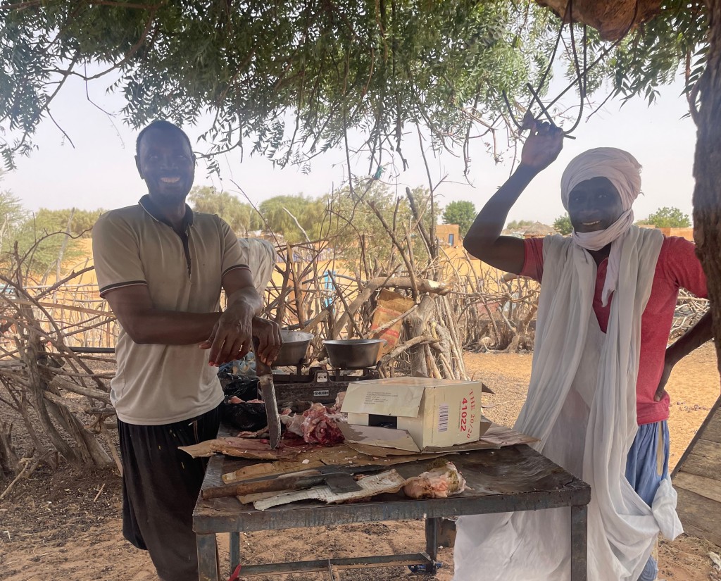 Two men prepare and weigh meat at an outdoor table shaded by a tree, with branches and a dry rural landscape in the background.