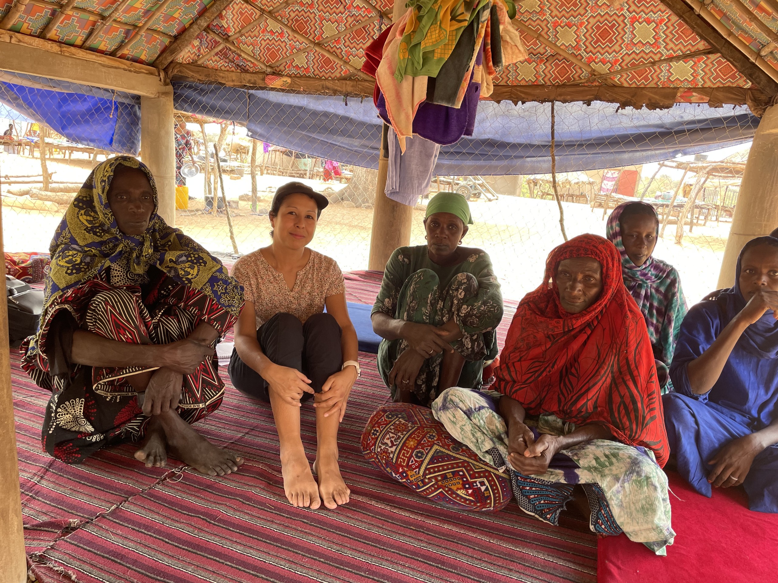 A group of women sit cross-legged on woven mats under a patterned fabric shelter, wearing colorful clothing and headscarves, gathered closely together in a shaded outdoor setting.