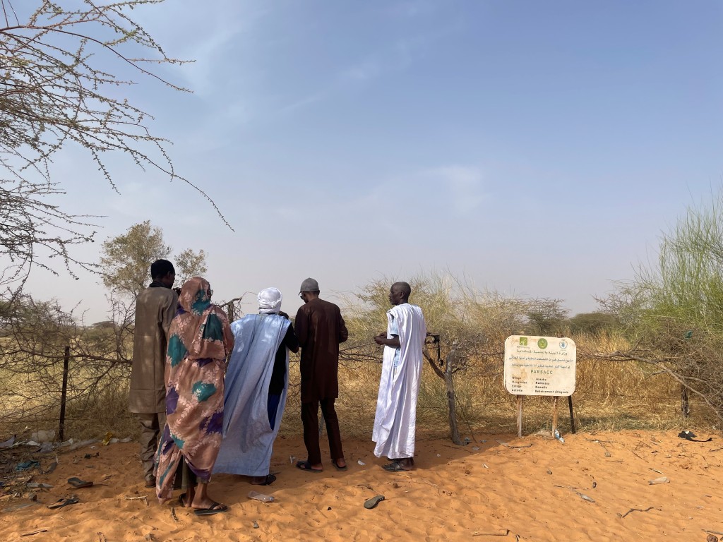 A small group of people stand on sandy ground beside a fenced plot with sparse shrubs and trees, gathered near a signboard in a dry, semi-arid landscape under a clear sky.